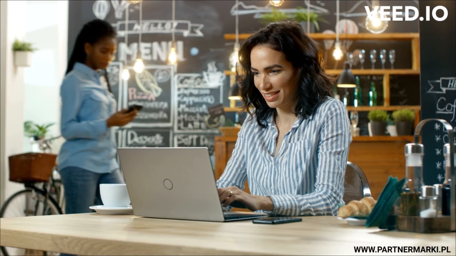 Woman working on laptop another woman in background using phone